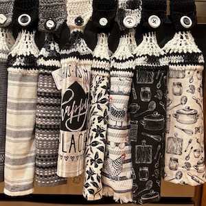 May include: A collection of hanging kitchen towels with crochet tops and button closures. The towels feature various patterns, including stripes, floral designs, and kitchen-themed illustrations in black, white, and gray. The text "THIS IS MY HAPPY PLACE" is visible on one towel.