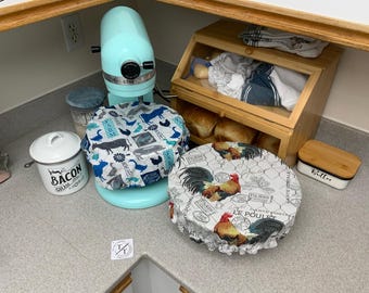 May include: A kitchen scene featuring a light blue stand mixer with a farm animal print cover. A white container labeled "Bacon Grease" sits nearby. A rooster print cover and a wooden bread box are also visible.