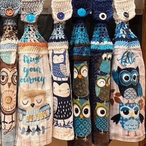May include: Six hanging kitchen towels with crocheted tops in shades of blue, white, and brown. Each towel features owl graphics and text including "trust your journey," "be awesome," and "hello." The tops have buttons for hanging on an oven handle.