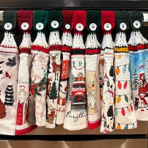 May include: A collection of hanging kitchen towels with crochet tops in red and green, each featuring a unique holiday design. Designs include snowmen, Santa, reindeer, and Christmas lights. The towels are attached to a metal bar.