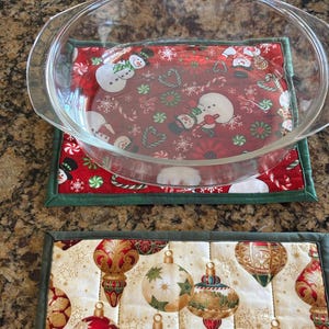 May include: A clear glass oval baking dish sits on a rectangular fabric mat featuring a red background with snowmen, candy canes, and snowflakes. Below it is a second rectangular fabric mat with a cream background and gold, red, and green ornaments.