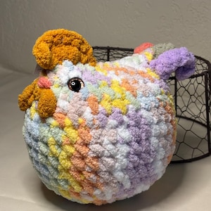 May include: A handmade, round crochet animal in pastel colors. The plush toy features a mix of yellow, orange, pink, purple, and blue yarn. It has brown eyes and orange accents on the head, resembling a chicken or bird.