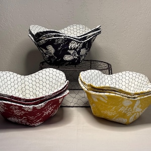 May include: A stack of fabric bowls in red, yellow, and navy blue, each with a unique pattern. The bowls have a white honeycomb pattern on the inside. The navy bowl has a rooster design, the red bowl has a floral design, and the yellow bowl has a floral design.