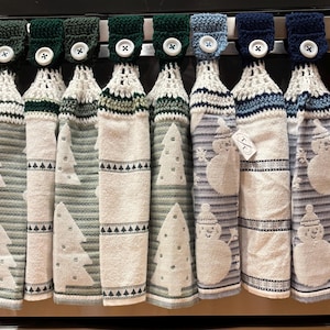 May include: Eight hanging kitchen towels with crocheted tops in shades of green and blue, each secured with a white button. The towels feature patterns of white Christmas trees or snowmen on striped backgrounds, designed to hang from a handle.