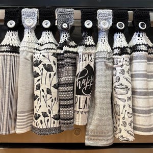 May include: A collection of hanging kitchen towels with crochet tops and button closures. The towels feature various patterns, including stripes, floral designs, and the text "THIS IS happy PLACE". The color palette is primarily black, white, and gray.