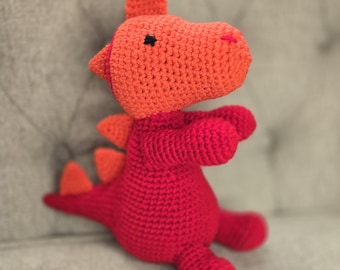 Può includere: Un giocattolo a forma di drago all'uncinetto fatto a mano in rosso e arancione vivace. Il drago presenta un design strutturato dettagliato con una testa appuntita, piccoli occhi neri e punte arancioni sulla schiena. Il giocattolo è morbido e soffice.