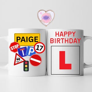 May include: Two white ceramic mugs. One mug has the name "PAIGE" in a yellow rectangle, surrounded by road signs. The other mug says "HAPPY BIRTHDAY" with a large red "L" inside a black square, a learner driver symbol.
