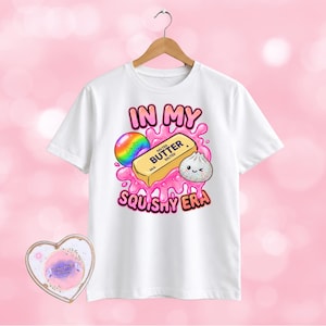 May include: A white short-sleeve t-shirt featuring a graphic print of a stick of butter, a rainbow sphere, and a small dumpling surrounded by pink liquid. The text reads "IN MY SQUISHY ERA". A heart-shaped card with a pink centre is positioned below.