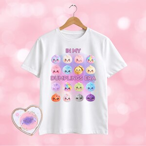 May include: A white short-sleeve t-shirt featuring a graphic print of sixteen colourful, cartoon-style dumplings with faces arranged in a grid. The text "IN MY DUMPLINGS ERA" is printed in pink above and below the grid of cute dumpling characters.