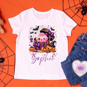 May include: A white short-sleeved t-shirt featuring a graphic print of a pink dumpling character inside a purple and orange gift box, surrounded by jack-o'-lanterns, bats, a spider web, and sweets. The name "Sophie" is printed in purple script below.