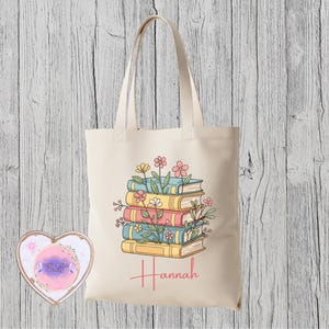 May include: A cream tote bag with a design of a stack of books in shades of yellow, pink, and blue, decorated with flowers. The name "Hannah" is written below the books in a cursive font. The bag has long handles.