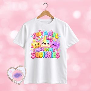 May include: A white short-sleeved t-shirt featuring a colourful graphic print of a rainbow-coloured character with a pink bow, a yellow butter block, and a purple cube, with the text "JUST A GIRL WHO LOVES SQUISHIES" in a bubble-letter font.