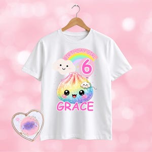 May include: A white short-sleeve t-shirt featuring a graphic of a rainbow-coloured dumpling with a smiling face, a pink balloon with the number 6, a rainbow, and two small clouds. The text reads BIRTHDAY GIRL and GRACE in pink lettering.