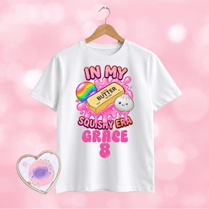 May include: A white short-sleeve t-shirt featuring a graphic print of a stick of salted butter, a rainbow sphere, and a small dumpling surrounded by pink liquid. The text reads IN MY SQUISHY ERA GRACE 8. A heart-shaped card is positioned below the shirt.
