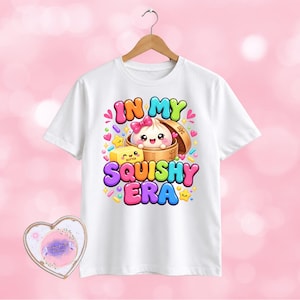 May include: A white t-shirt featuring a colourful graphic of a smiling dumpling in a bamboo steamer and a block of butter with the text IN MY SQUISHY ERA, surrounded by small hearts, stars, and sprinkles, hanging on a wooden hanger.