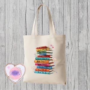 May include: A cream tote bag displaying a stack of colourful books with titles and floral accents. A heart-shaped coaster with a watercolour design is on the left.