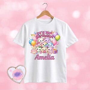May include: A white short-sleeved t-shirt featuring a colourful graphic of cartoon dumplings, balloons, a birthday cake, and a gift box. The text reads It's My Birthday and the name Amelia in pink lettering. The shirt hangs on a wooden hanger.