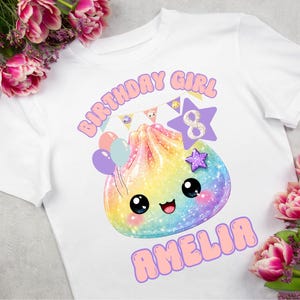 May include: A white t-shirt featuring a colourful, glittery, rainbow-hued dumpling character with a smiling face. The graphic includes the text "BIRTHDAY GIRL" and "AMELIA," a purple star with the number "8," and a string of small pennant flags.