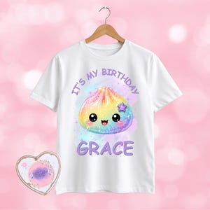 May include: A white short-sleeved t-shirt featuring a graphic of a rainbow-coloured dumpling with a smiling face, a small purple star, and the text "IT'S MY BIRTHDAY GRACE" in purple lettering. A heart-shaped tag with matching graphics is included.
