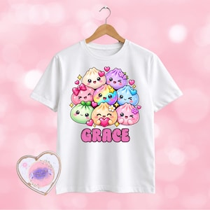 May include: A white short-sleeved t-shirt featuring a graphic of multiple colourful, smiling dumpling characters with hearts and flowers. The name GRACE is printed in large pink bubble letters below the dumplings. A heart-shaped tag is visible.