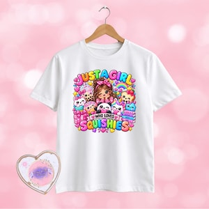 May include: A white short-sleeve t-shirt featuring a colourful graphic print of a cartoon character surrounded by various squishy toys, boba tea, and hearts. The text reads JUST A GIRL WHO LOVES SQUISHIES. A heart-shaped decorative item is nearby.