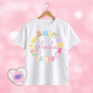 May include: A white short-sleeved t-shirt featuring a circular arrangement of colourful cartoon icons including a banana, frog, duck, and toast, surrounding the name Amelia written in pink script. The shirt hangs on a wooden hanger.