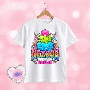 May include: A white short-sleeved t-shirt featuring a colourful graphic of a green cat sitting on a blue blob with a pink spiky ball on top. The text NEEDOH and AMELIA appear in a stylised, dripping font below the cat illustration.