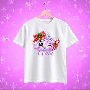 May include: A white short-sleeved t-shirt featuring a graphic of a purple dumpling character with a red bow, holly, and a candy cane. The character has large eyes and a smiling mouth. The name Grace is printed in purple text below the graphic.