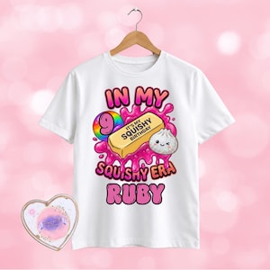 May include: A white t-shirt featuring a graphic with the text IN MY SQUISHY ERA RUBY and a number 9. The design includes a yellow rectangular squishy toy with the text IT'S MY SQUISHY BIRTHDAY, a small white dumpling character, and pink slime.