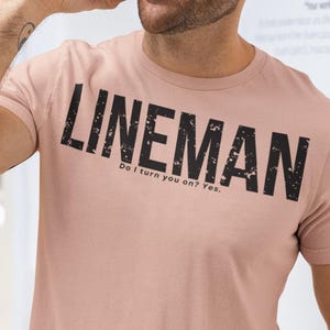 Op de afbeelding: Een oudroze T-shirt met korte mouwen en een ronde hals, voorzien van het woord LINEMAN in grote, verweerde zwarte hoofdletters op de borst, met de kleinere tekst Do I turn you on? Yes. direct onder het hoofdwort gedrukt.
