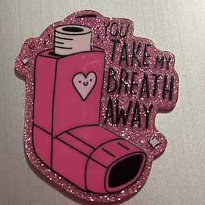 May include: A pink glittery pin shaped like an inhaler with the text "YOU TAKE MY BREATH AWAY". The inhaler has a white top and a heart with a smiling face. The pin is on a silver surface.