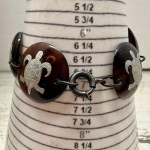 May include: A brown and silver bracelet featuring a turtle design. The bracelet has oval-shaped links with a silver turtle design on each link. The bracelet is connected with a silver clasp. The bracelet is approximately 7 inches in length.