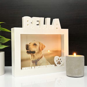 Könnte beinhalten: Weißer Holzrahmen mit einem Foto eines Hundes und dem Namen "BELLA" oben. Der Rahmen hat ein kleines Herz mit einem Pfotenabdruck darin und den Text "IMMER IN UNSEREN HERZEN" unten. Ein kleiner Beton-Kerzenhalter mit einer brennenden Kerze steht neben dem Rahmen.
