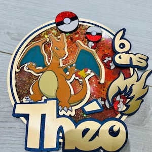 May include: A gold and blue cake topper featuring a cartoon image of a red and orange Pokemon character, a red and white Pokeball, and the text "Theo 6 ans".