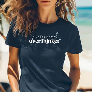 May include: Navy blue t-shirt with the white text "professional overthinker" in a cursive font. The shirt has short sleeves and a crew neck. The background is a blurred image of a beach.