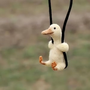 May include: A small, white, felt duck is hanging from a black string. The duck has a small, orange beak and orange feet. The duck is sitting on a swing.