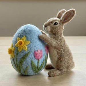 May include: A felt Easter bunny figurine beside a light blue egg decorated with yellow daffodils and pink tulips. The brown bunny has long ears and is leaning against the egg, a springtime theme.