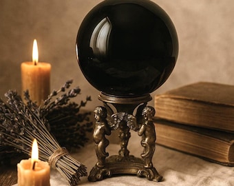 May include: A black crystal ball sits on a gold metal stand with two cherub figures. The stand is on a linen cloth with two lit candles and dried lavender.