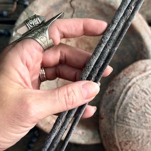 Peut inclure: Trois bâtonnets d'encens gris foncé sont tenus dans une main. Les bâtonnets d'encens ont un aspect texturé. Une grande bague en argent avec un cristal clair est portée au doigt. L'arrière-plan comprend une surface rustique et une assiette décorative.