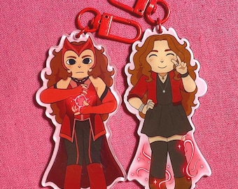 May include: Two acrylic keychains featuring cartoon illustrations of a character in red and black outfits. The keychains have red lobster clasp closures. The background is a solid pink color.