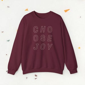 May include: A burgundy sweatshirt featuring the white text "CHOOSE JOY", arranged vertically. The crew neck sweatshirt has long sleeves. The text is in a clean, sans-serif font.