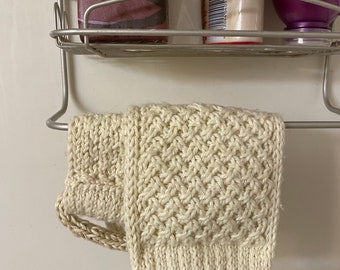 May include: A close-up of a beige knitted bath pouf with a loop handle. The pouf is hanging on a metal rack in a bathroom.