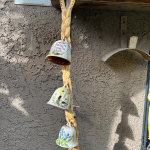 May include: A set of three ceramic bells with floral designs hanging from a woven rope. The bells are white with blue, yellow, and purple flowers painted on them.