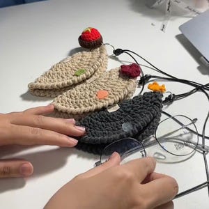 May include: Crocheted eyeglass cases in beige and charcoal gray, each with a decorative charm. The cases have a button closure and a black cord. One case has a red strawberry charm, another has a red heart, and another has an orange fish.