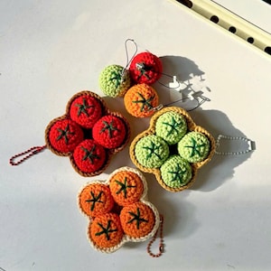 Może przedstawiać: Szydełkowe breloczki do kluczy z motywem pomidorów w kolorze czerwonym, zielonym i pomarańczowym. Każdy breloczek przedstawia grupę szydełkowych pomidorów z zielonymi łodygami. Breloczki są w różnych kolorach i stylach, idealne do dodania odrobiny fantazji.