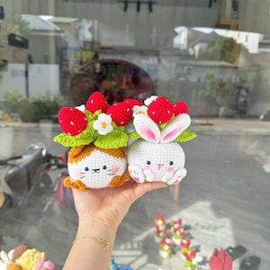 May include: Two crocheted amigurumi figures: a white bunny and a cat with orange and white fur. Both have strawberry and leaf accents on their heads. The bunny has pink inner ears and paws. The cat has a small orange nose and whiskers.