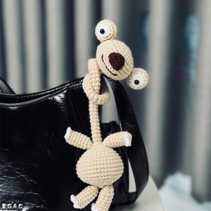 Pode incluir: Uma criatura bege de crochê com olhos grandes e arregalados e um nariz em forma de coração marrom, pendurada em uma bolsa preta. O brinquedo de pelúcia feito à mão tem um pescoço longo e braços e pernas pequenos com pontas brancas.