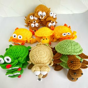 Puede incluir: Una colección de criaturas de ganchillo hechas a mano, que incluyen cangrejos, tortugas y otros diseños caprichosos. Las criaturas son de varios colores, como amarillo, verde y marrón, con acentos blancos y negros. La colección se exhibe sobre una superficie blanca.