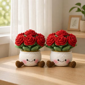 Pode incluir: Duas floreiras de crochê feitas à mão, cada uma com um rosto sorridente, segurando um buquê de rosas vermelhas. Os vasos são brancos com folhas verdes e bases amarelas. Acentos em forma de coração marrom são colocados ao redor dos vasos.