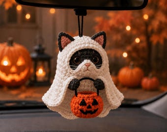 Pode incluir: Um ornamento de gato em crochê pendurado num espelho retrovisor. O gato preto e branco veste um traje de fantasma branco com capuz e segura um pequeno balde de crochê laranja em forma de abóbora com um rosto preto desenhado.